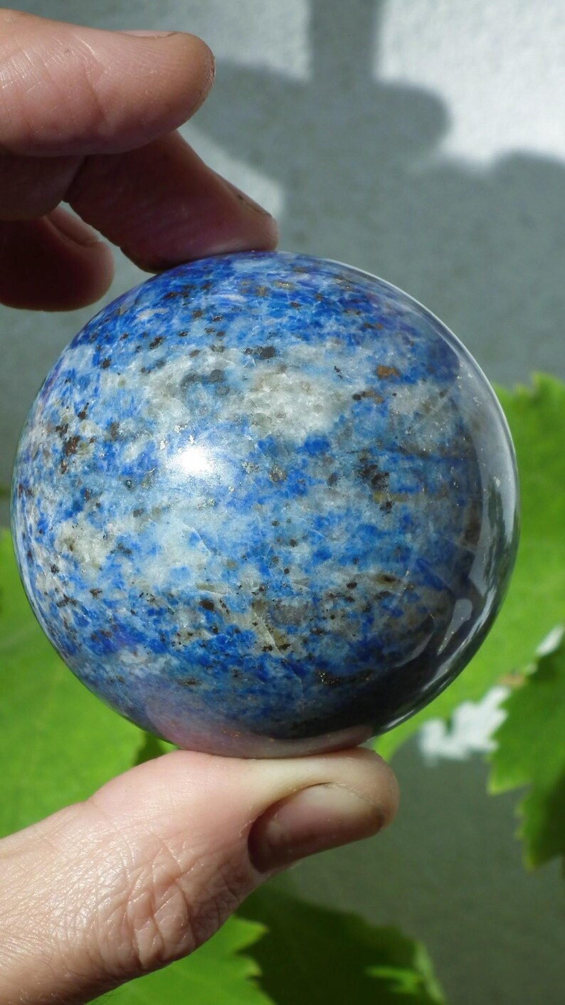 May include: A polished, spherical lapis lazuli gemstone. The stone is a deep blue color with white and gold flecks throughout. The sphere is held in a hand, with a green leafy background.