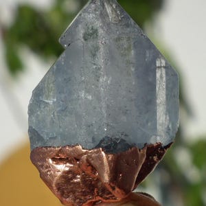 K&ouml;nnte beinhalten: Ein durchscheinender, hellblauer Kristallpunkt mit einer gezackten, texturierten kupferfarbenen Metallbasis. Der Kristall weist nat&uuml;rliche Facetten und Einschl&uuml;sse auf, w&auml;hrend der metallische Boden eine raue, organische Oberfl&auml;che hat.