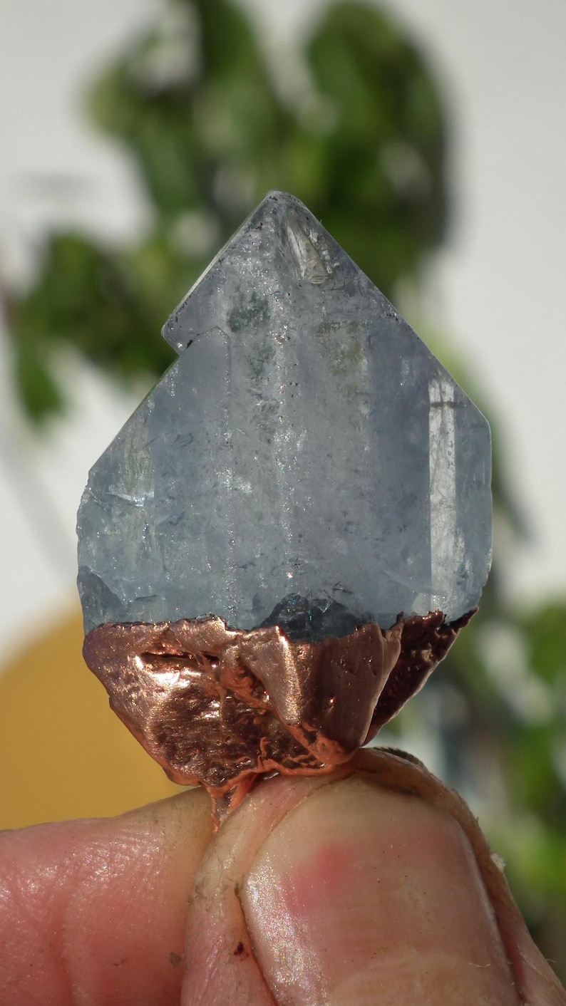 K&ouml;nnte beinhalten: Ein durchscheinender, hellblauer Kristallpunkt mit einer gezackten, texturierten kupferfarbenen Metallbasis. Der Kristall weist nat&uuml;rliche Facetten und Einschl&uuml;sse auf, w&auml;hrend der metallische Boden eine raue, organische Oberfl&auml;che hat.