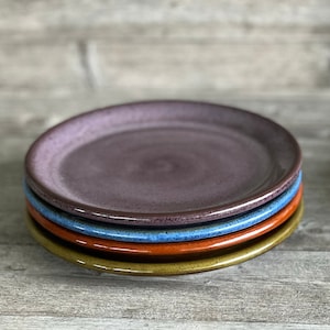 May include: Four ceramic plates stacked on top of each other. The top plate is purple, the next is blue, the next is orange, and the bottom plate is green.