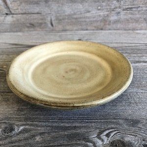 May include: A small, round, light yellow ceramic plate with a slightly raised rim. The plate has a subtle swirl pattern and a speckled glaze.