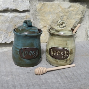 May include: Two ceramic honey pots with lids, one blue and one beige, with the word "honey" inscribed on each. A wooden honey dipper is in the foreground.