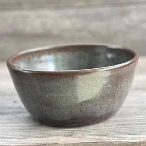 May include: A small, round ceramic bowl with a speckled brown and gray glaze. The bowl has a slightly uneven rim and a smooth, glossy finish.