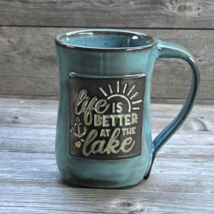 May include: A teal blue ceramic mug with a black square panel that reads "life is better at the lake" in gold lettering. The panel also features a sun, anchor, and heart.