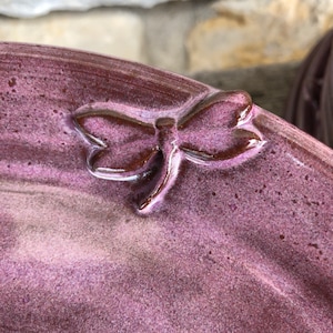 May include: A purple ceramic dish with a raised dragonfly design. The dragonfly is brown and has a glossy finish.