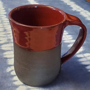 May include: A red and black ceramic mug with a rounded handle. The mug has a dark brown glaze on the bottom half and a glossy red glaze on the top half.