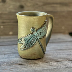 May include: A green ceramic mug with a dragonfly design. The dragonfly is blue and green and has a raised texture.