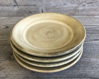 May include: A stack of four ceramic plates in a light beige color with a speckled glaze. The plates have a slightly raised rim and a subtle swirl pattern in the center.