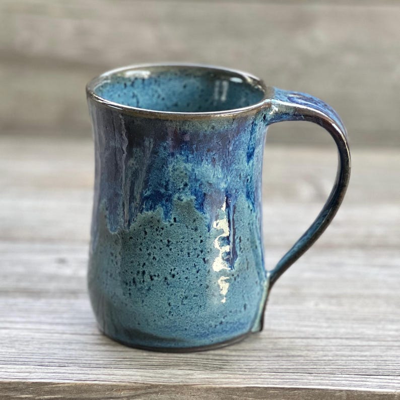 May include: A handmade ceramic mug with a blue and green glaze. The mug has a wide, rounded body and a comfortable handle.