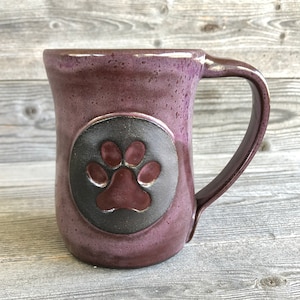May include: Purple ceramic mug with a black circle and a raised paw print in the center. The mug has a rounded handle.
