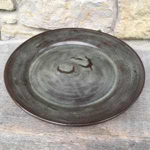 May include: A large, round, ceramic plate with a dark brown glaze and a green swirl pattern. The plate has a slightly raised rim.