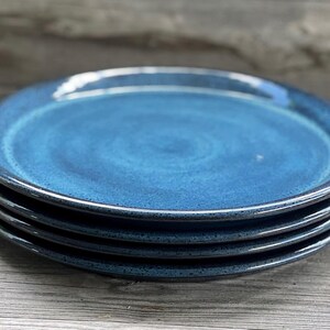 May include: A stack of four blue ceramic plates with a speckled texture and a dark rim. The plates are a deep shade of blue, with a glossy finish. The plates are stacked on a wooden surface.