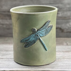 May include: A sage green ceramic tumbler with a raised teal dragonfly design. The cylindrical cup has a slightly flared rim and a smooth, matte finish. The dragonfly is detailed with textured wings.