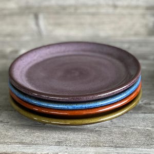 May include: Four ceramic plates stacked on top of each other. The top plate is a deep purple color, the next plate is a light blue color, the next plate is a bright orange color, and the bottom plate is a light yellow color.
