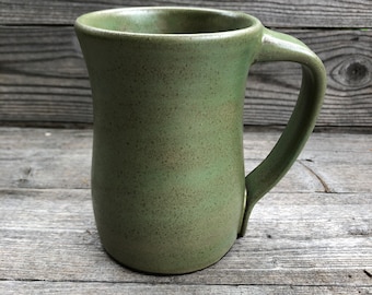 May include: A green ceramic mug with a speckled glaze. The mug has a rounded body and a wide handle.