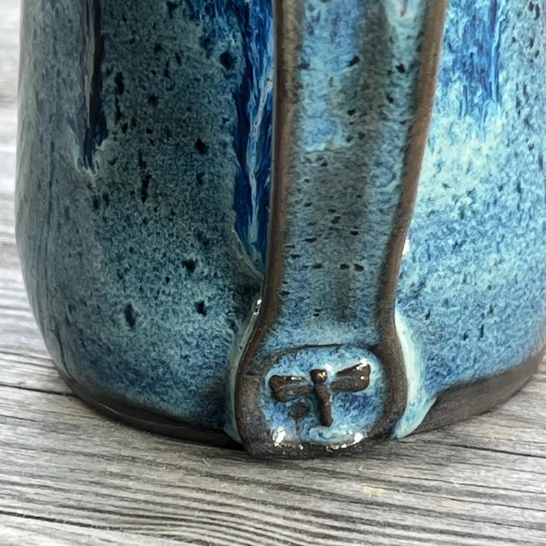 May include: A blue ceramic mug with a dragonfly design on the handle. The mug has a speckled glaze and a dark blue color.