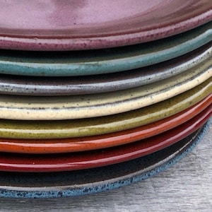 May include: A stack of handmade ceramic plates in various colors, including deep red, teal, and yellow. The plates have a glossy finish and a slightly textured rim. The plates are stacked on top of each other, showcasing their different colors and textures.