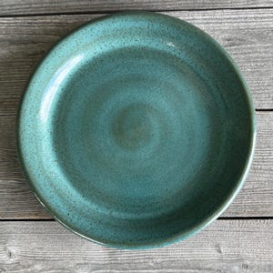 May include: A teal ceramic dinner plate with a speckled glaze. The plate has a slightly raised rim and a smooth, glossy finish.