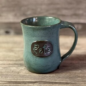 May include: A teal blue ceramic mug with a brown oval label that says "Pete".