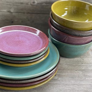 May include: A stack of five ceramic plates in shades of teal, yellow, brown, and pink. The plates are stacked on a wooden surface.
