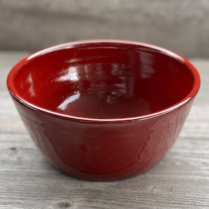 May include: A deep red ceramic bowl with a glossy finish. The bowl is round and has a slight lip around the rim.