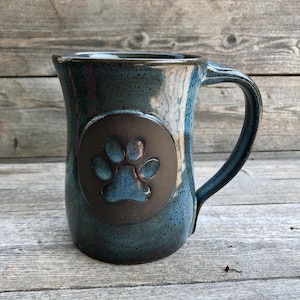 May include: A blue ceramic mug with a brown paw print design. The mug has a rounded body and a handle.