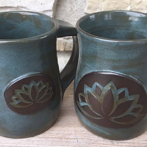 May include: Two blue ceramic mugs with brown lotus flower designs. The mugs have a rounded shape and a handle on the side.