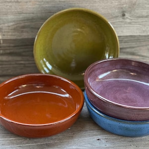 May include: A collection of handmade ceramic bowls in various colors. The bowls are in shades of orange, olive green, purple, and blue. The bowls have a glossy finish and are stacked on a wooden surface.