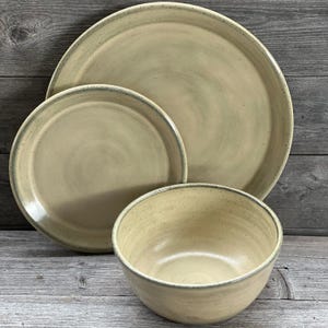 May include: A set of handmade ceramic dinnerware in a muted yellow-green hue. The set includes a large plate, a smaller plate, and a bowl. The plates and bowl have a speckled texture and a darker rim.