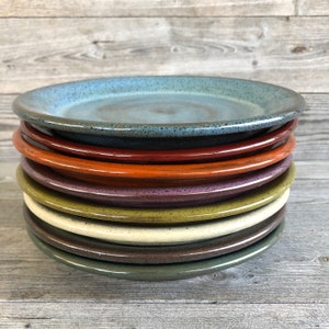 May include: A stack of eight ceramic plates in various colors, including blue, red, orange, purple, green, beige, and brown. The plates are all round and have a slightly textured surface.