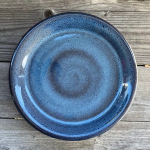 May include: A blue ceramic plate with a speckled glaze. The plate has a slightly raised rim and a subtle swirl pattern.