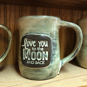 May include: A light green ceramic mug with a brown square on the front that says "Love you to the moon and back".