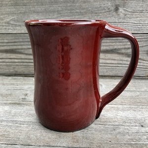 May include: A deep red ceramic mug with a rounded body and a handle. The mug has a smooth, glossy finish.