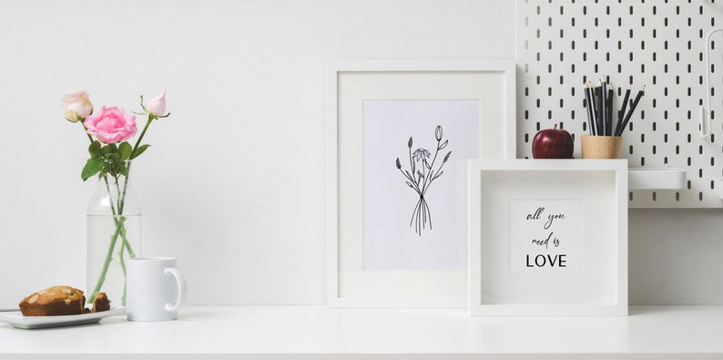 May include: White framed artwork featuring a black line drawing of flowers. A white framed print with the text "all you need is LOVE" sits on a white surface with a vase of pink roses, a white mug, and a small plate with a muffin.