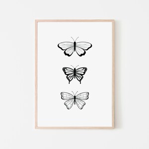 May include: A framed print featuring three stylized butterfly illustrations in black and white. Each butterfly has a unique wing pattern, with the artwork set against a white background. The frame is a light wood color.