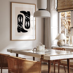May include: A black and white illustration of cowboy boots hangs on a wall above a white dining table with a glass top. Two wicker chairs are positioned on either side of the table. A white ceramic lamp sits on a table next to a window with a bamboo blind.