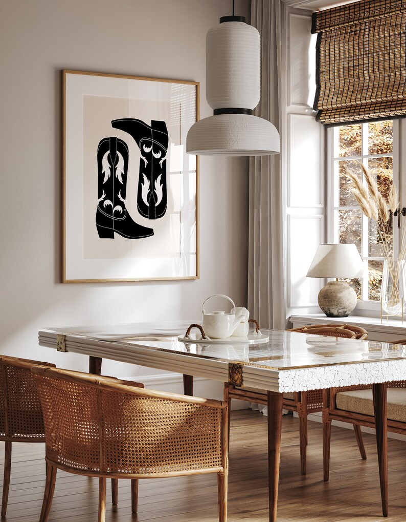 May include: A black and white illustration of cowboy boots hangs on a wall above a white dining table with a glass top. Two wicker chairs are positioned on either side of the table. A white ceramic lamp sits on a table next to a window with a bamboo blind.