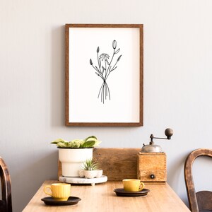May include: A framed black and white line drawing of a bouquet of flowers, hanging on a white wall above a wooden table with two yellow mugs and a wooden coffee grinder.