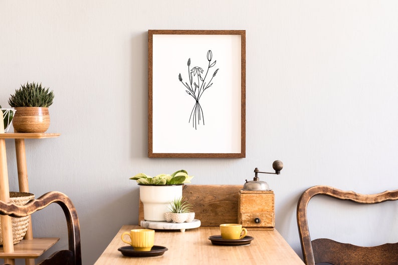 May include: A framed black and white line drawing of a bouquet of flowers, hanging on a white wall above a wooden table with two yellow mugs and a wooden coffee grinder.