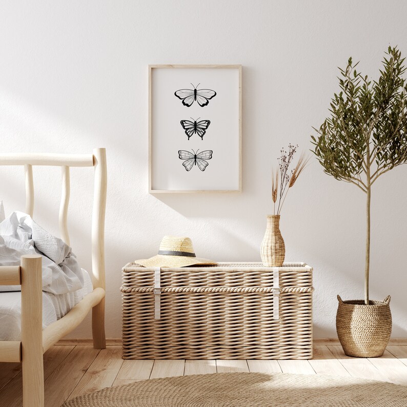 May include: A framed print featuring three black and white butterfly illustrations. The artwork hangs above a woven wicker trunk with a straw hat on top. A small vase with dried plants and a potted tree complete the scene.