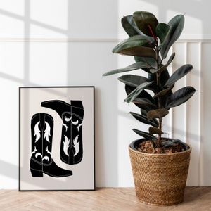 May include: Black and white illustration of two cowboy boots with a white design on the front. The illustration is framed and hanging on a white wall next to a potted plant.