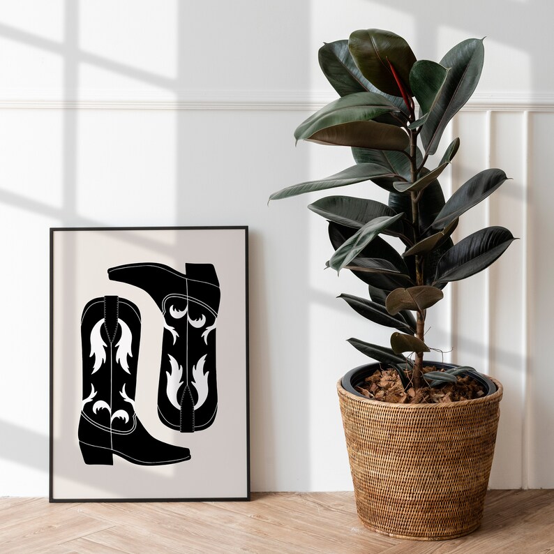 May include: Black and white illustration of two cowboy boots with a white design on the front. The illustration is framed and hanging on a white wall next to a potted plant.