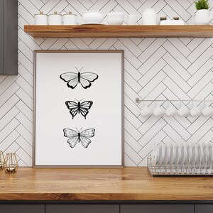 May include: A framed art print featuring three black and white butterfly illustrations. The artwork is displayed on a wooden countertop in a kitchen setting, with a white tile backsplash and a shelf with kitchenware.