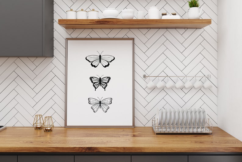 May include: A framed art print featuring three black and white butterfly illustrations. The artwork is displayed on a wooden countertop in a kitchen setting, with a white tile backsplash and a shelf with kitchenware.