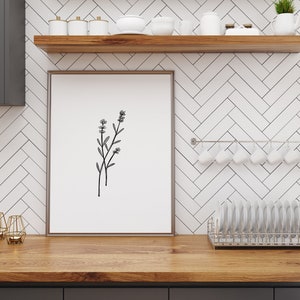 May include: A framed black and white line drawing of a sprig of flowers hangs on a white subway tile wall in a kitchen. The wall has a wooden shelf above the artwork and a metal rack with white mugs hanging below the artwork.