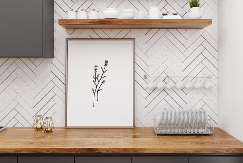 May include: A framed black and white line drawing of a sprig of flowers hangs on a white subway tile wall in a kitchen. The wall has a wooden shelf above the artwork and a metal rack with white mugs hanging below the artwork.