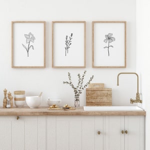 May include: Three framed black and white line drawings of flowers hanging on a white wall above a wooden countertop with a sink and faucet. The drawings are of a daffodil, a sprig of lavender, and a single flower.