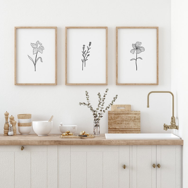 May include: Three framed black and white line drawings of flowers hanging on a white wall above a wooden countertop with a sink and faucet. The drawings are of a daffodil, a sprig of lavender, and a single flower.