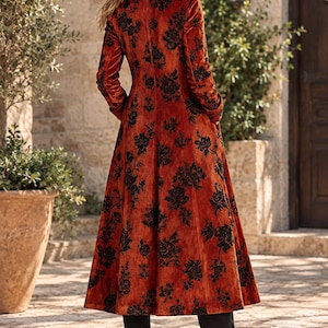 May include: A long, burnt-orange velvet coat featuring a black floral pattern, a structured collar, and long sleeves. The coat is worn over black trousers and black block-heeled boots, extending to the mid-calf with a flared, flowing silhouette.