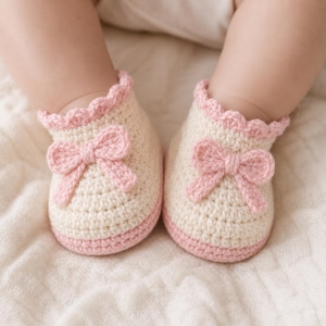 May include: A pair of cream-colored crochet baby booties featuring a soft pink scalloped trim at the ankle and a matching pink crochet bow centered on the front of each shoe. The booties have a solid pink base and a textured, hand-stitched appearance.